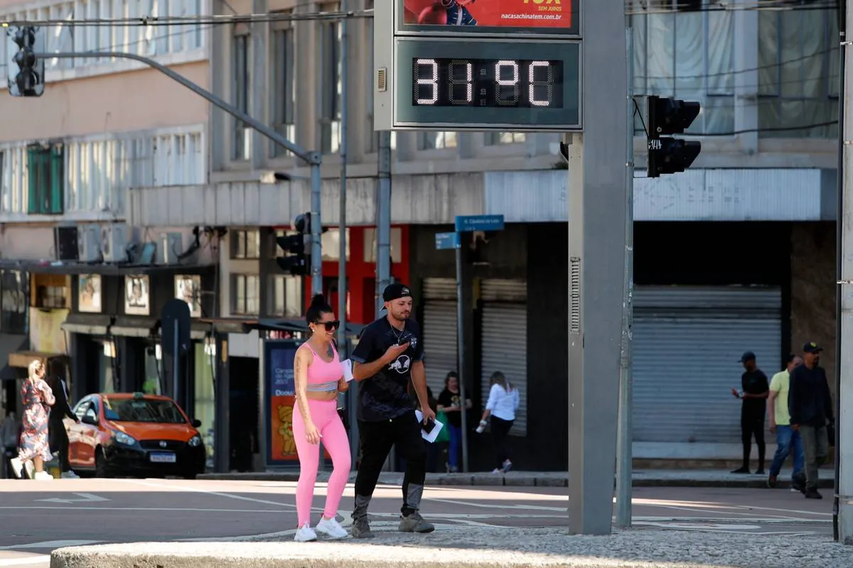 Imagem mostra o centro de Curitiba, com um casal andando em frente a um termômetro de rua com a temperatura de 31ºC