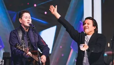 Imagem mostra a dupla se apresentando, com gian tocando um violão e giovani acenando para o público segurando um microfone