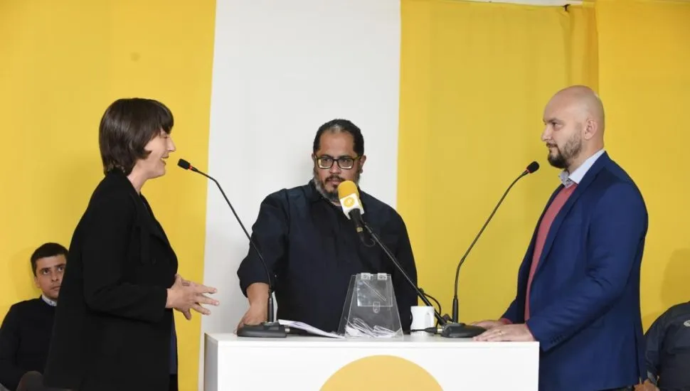 Na imagem, candidatos falam ao microfone, um em frente ao outro. Mediador do debate está entre eles.