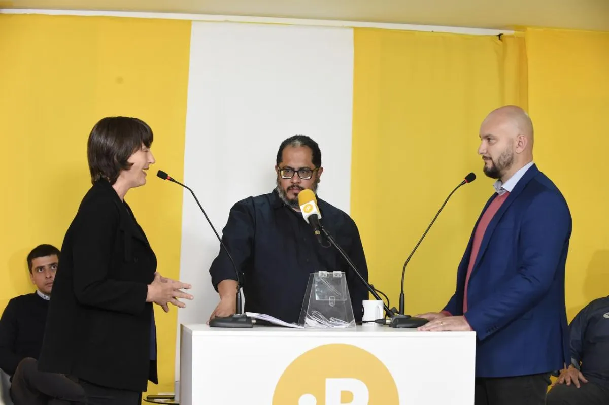 Na imagem, candidatos falam ao microfone, um em frente ao outro. Mediador do debate está entre eles.