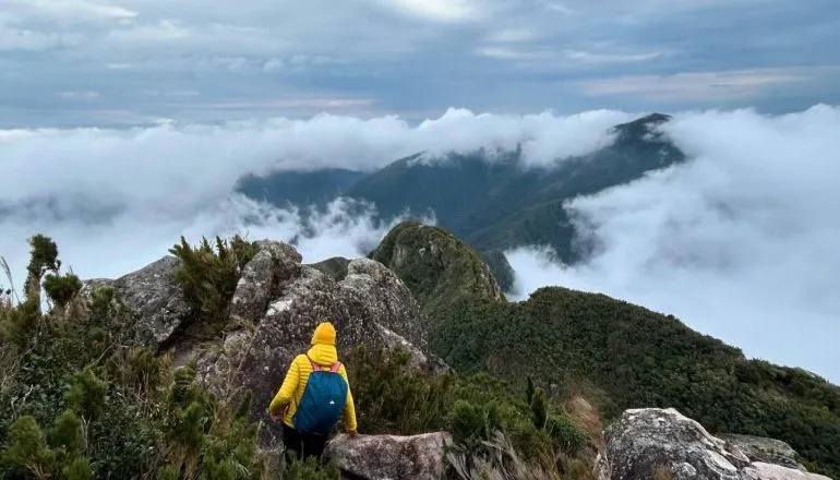 Na imagem, umas pessoa faz trilha nas montanhas. Paisagem tem nuvens sobre os morros.