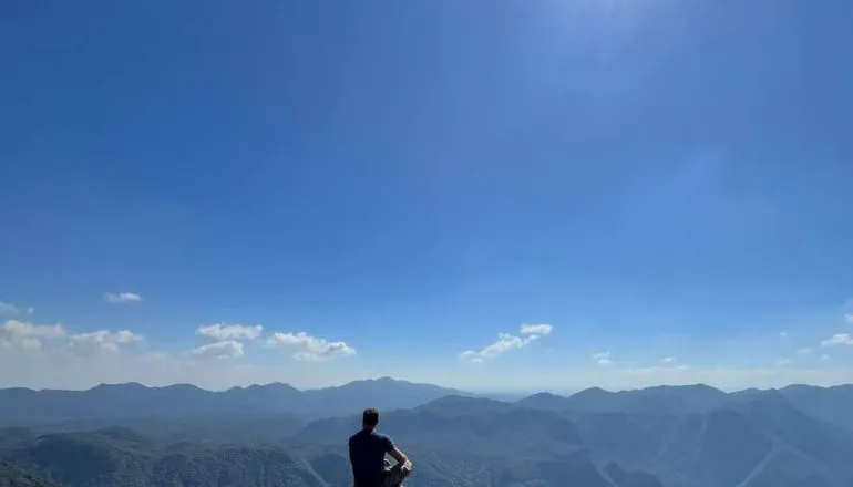 Na imagem, homem sentado em uma pedra contempla as montanhas.