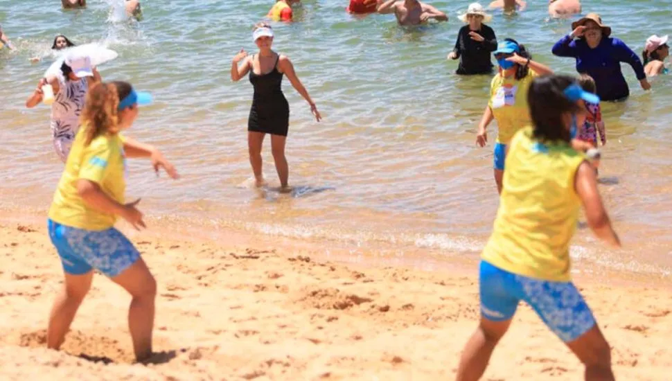 Na imagem, pessoas dançam na praia, acompanhando a coreografia de duas professoras.