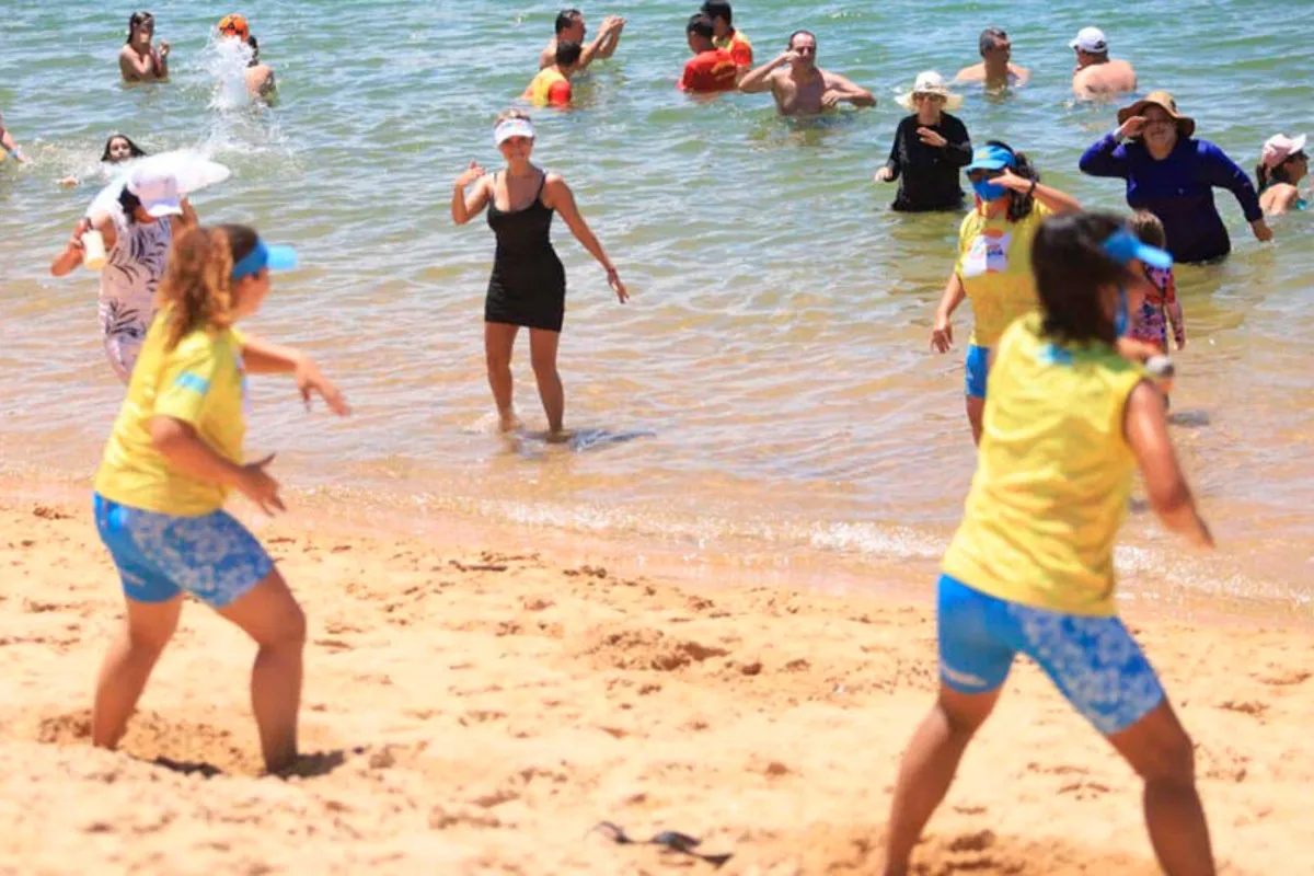 Na imagem, pessoas dançam na praia, acompanhando a coreografia de duas professoras.