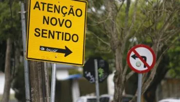 Na imagem, placas de rua indicam novo sentido e proibido virar à esquerda.