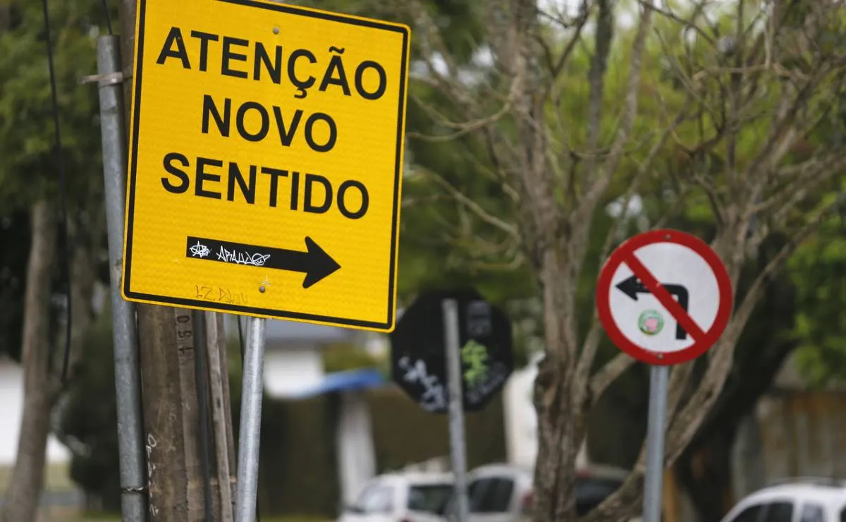 Na imagem, placas de rua indicam novo sentido e proibido virar à esquerda.