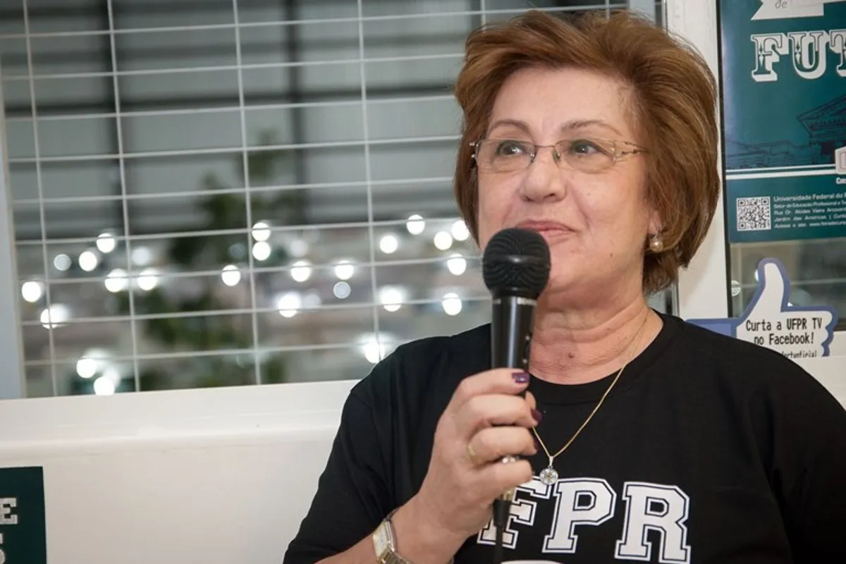 Na imagem, a professora Maria Amélia segura um microfone. Ela veste uma camiseta com a sigla da UFPR.