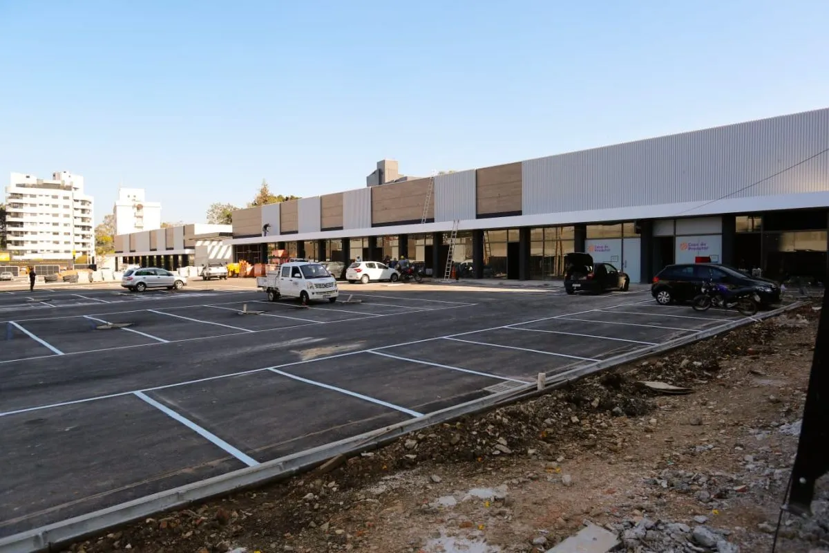 Imagem mostra algumas das lojas do novo shopping, com vagas de estacionamentos e alguns carros parados. Ao fundo, prédios e na frente da imagem restos de obras