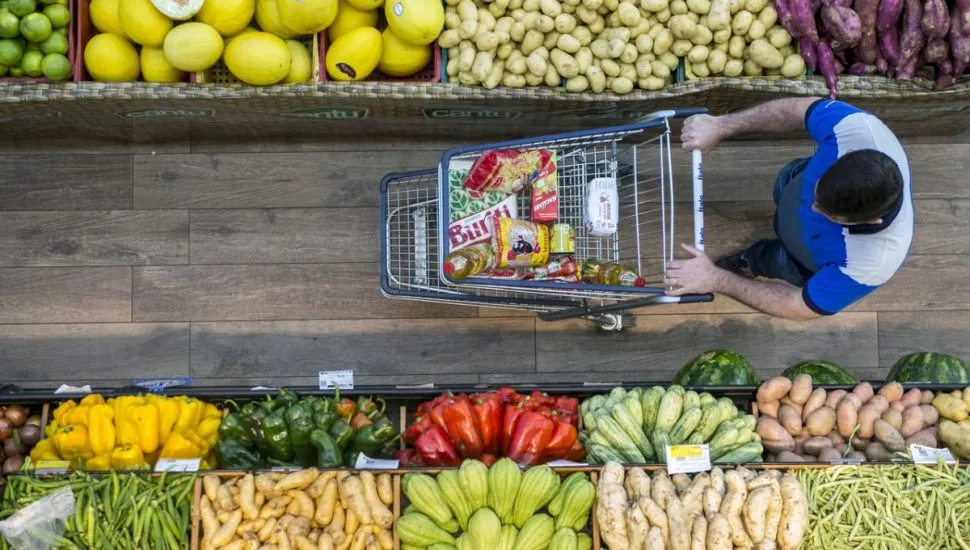 Na imagem, um homem empurra um carrinho com itens de suas compras, no corredor de frutas e verduras do supermercado.