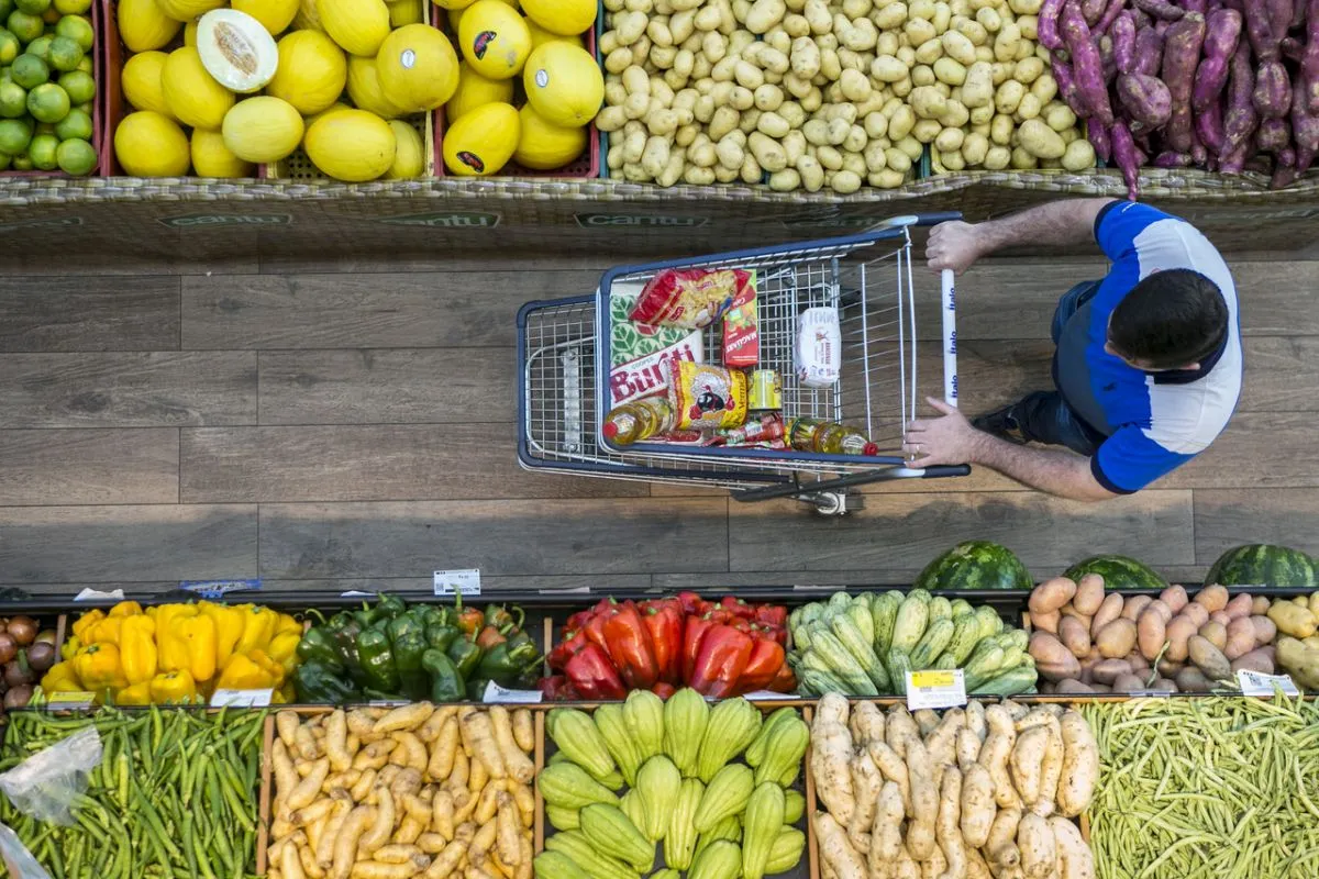 Na imagem, um homem empurra um carrinho com itens de suas compras, no corredor de frutas e verduras do supermercado.