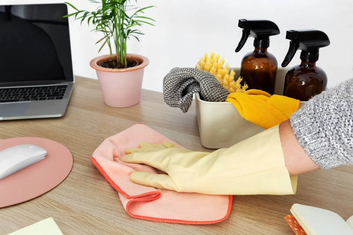 Imagem mostra uma pessoa passando um pano de limpeza em uma mesa com um computador e dois borrifadores.