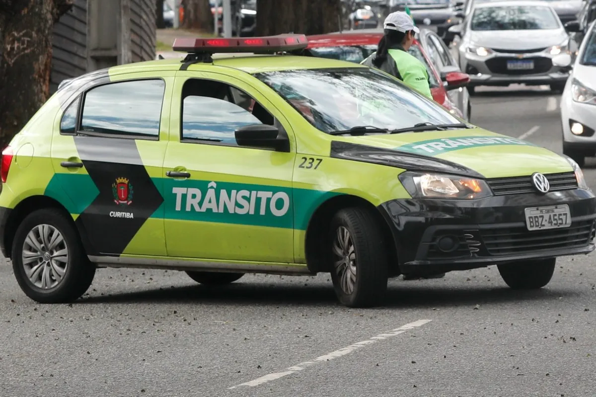 Imagem mostra uma viatura da Setran, parada e bloqueando uma faixa de uma rua. Há uma agente de trânsito atrás do carro, de costas.