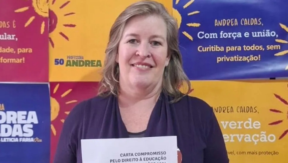 candidata Professora Andrea Caldas está em frente a um banner segurando uma carta de compromissos para a educação.