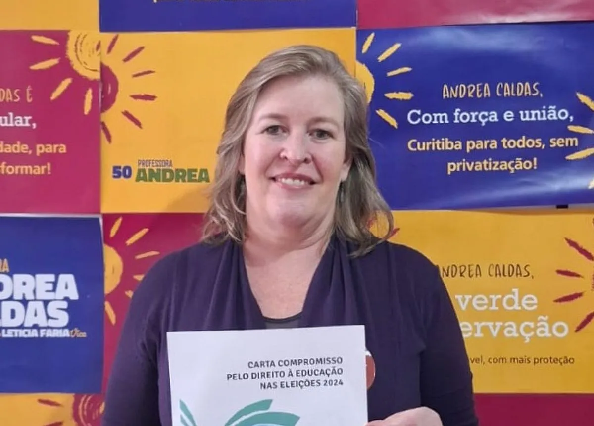 candidata Professora Andrea Caldas está em frente a um banner segurando uma carta de compromissos para a educação.