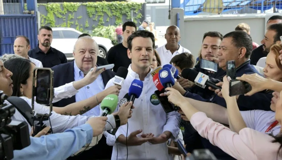 Na imagem, Eduardo Pimentel dá entrevista a jornalistas, acompanhado de Rafael Greca. Ele fala diante de muitos microfones da imprensa.