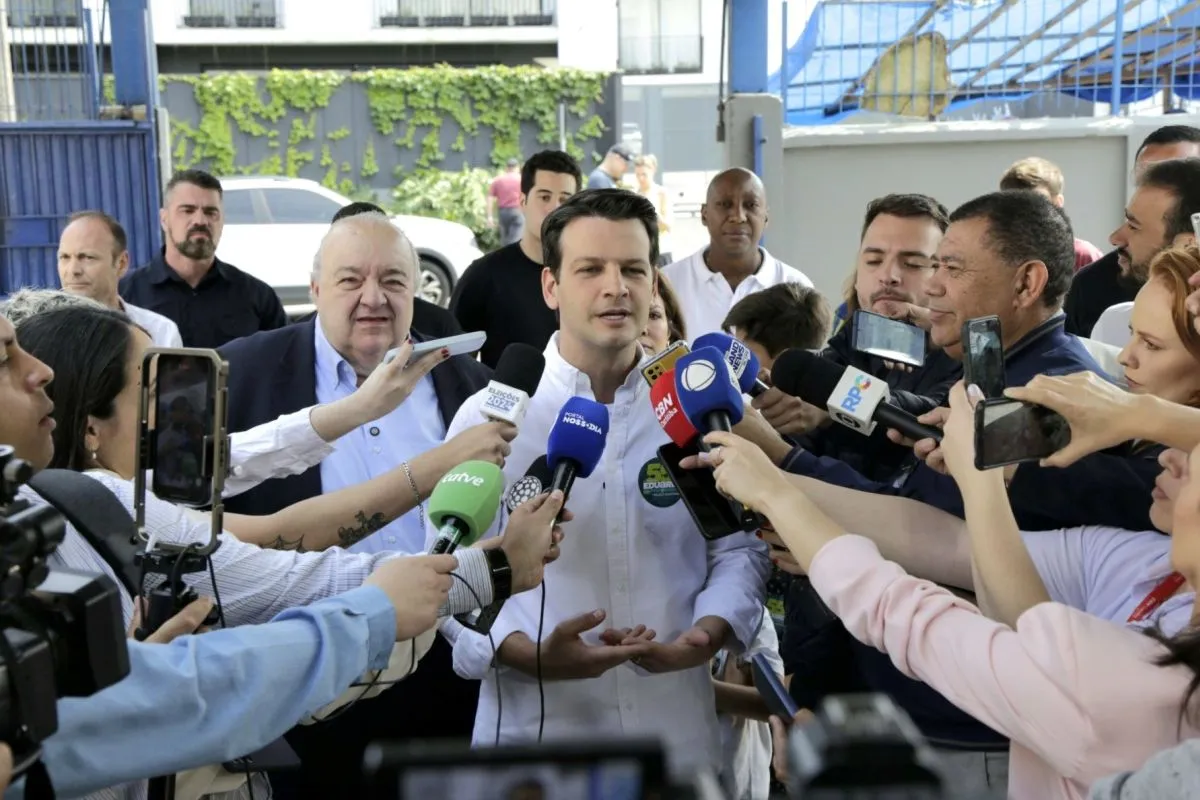 Na imagem, Eduardo Pimentel dá entrevista a jornalistas, acompanhado de Rafael Greca. Ele fala diante de muitos microfones da imprensa.