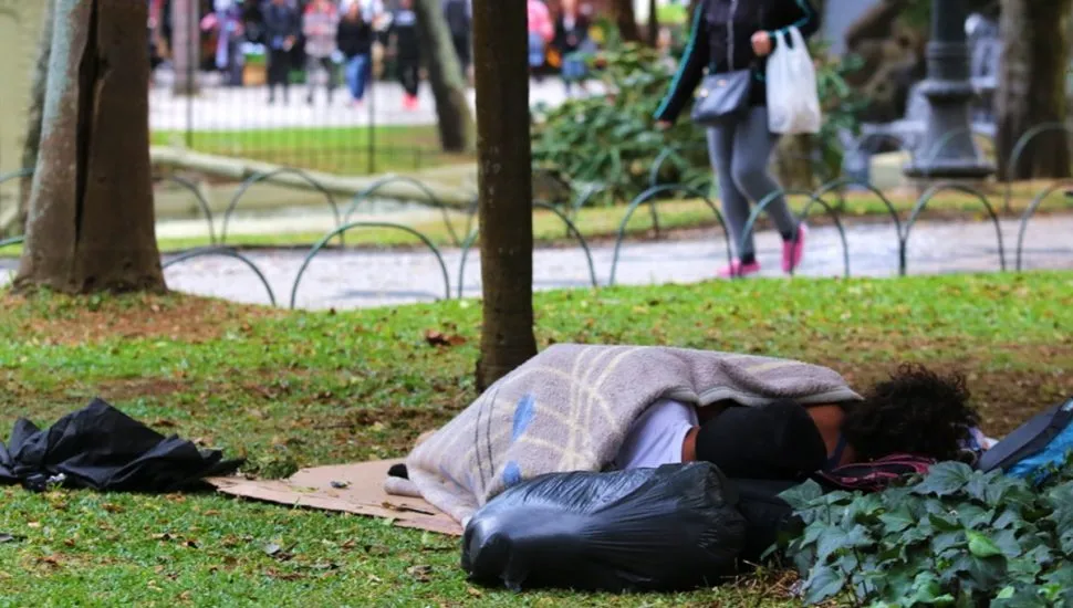 Na imagem, duas pessoas em situação de rua dormem no gramado de uma praça.