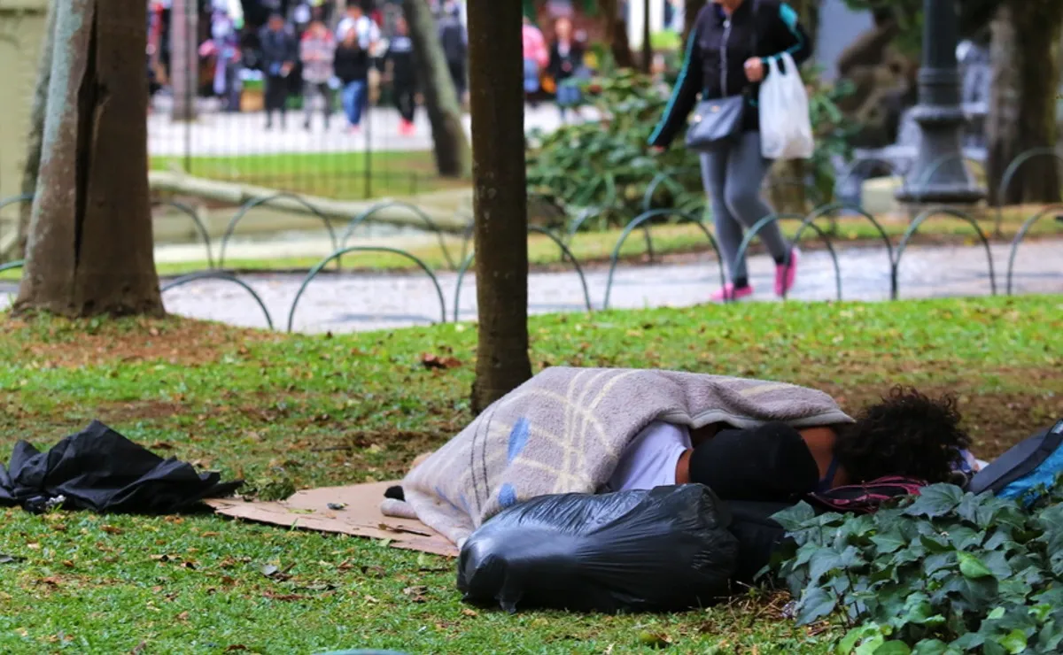 Na imagem, duas pessoas em situação de rua dormem no gramado de uma praça.