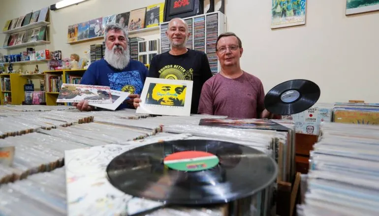 Imagem mostra um trio de amigos entre milhares de discos de vinil.