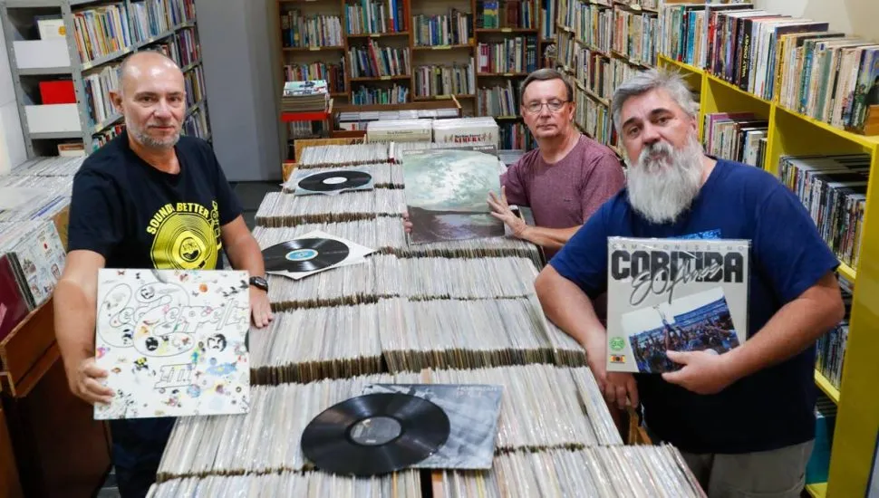 Imagem mostra um trio de amigos entre milhares de discos de vinil.