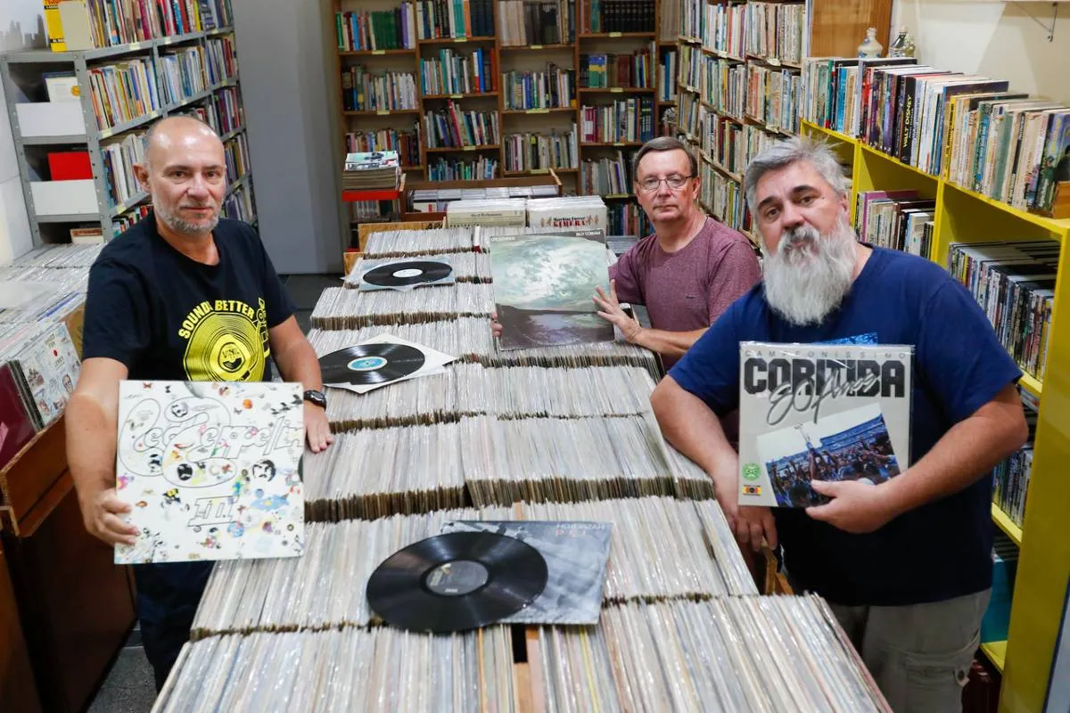 Imagem mostra um trio de amigos entre milhares de discos de vinil.