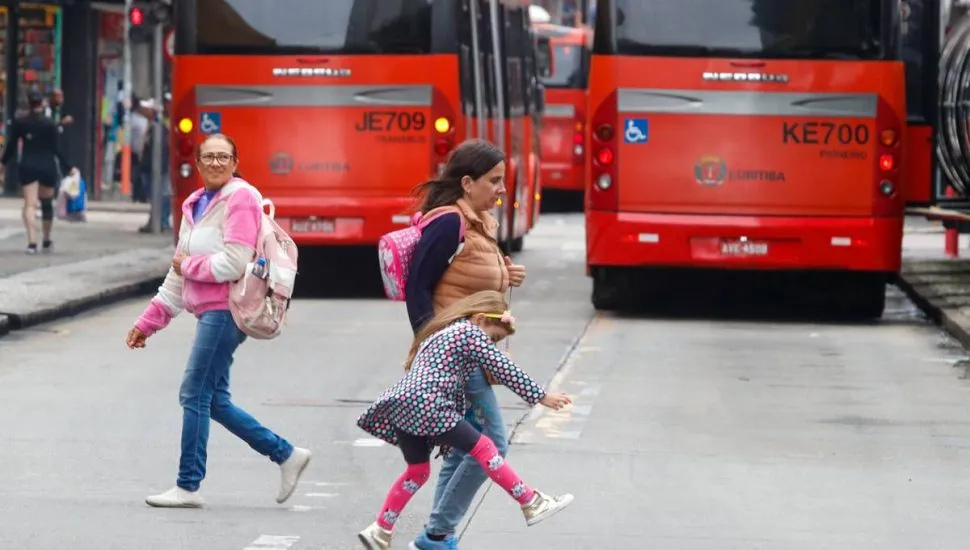 Imagem mostra dois biarticulados em Curitiba com pedestres atravessando a rua. Um dos pedestres é uma criança travessa.