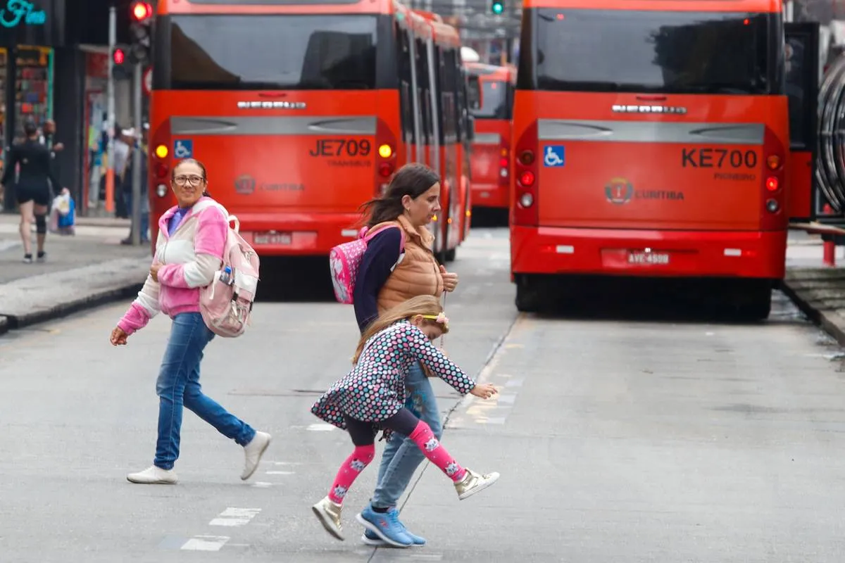 Imagem mostra dois biarticulados em Curitiba com pedestres atravessando a rua. Um dos pedestres é uma criança travessa.