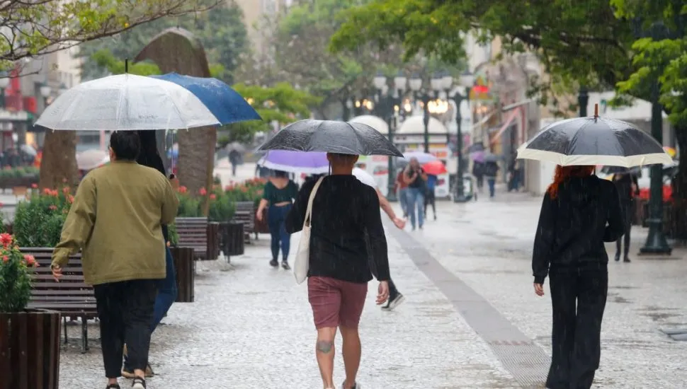 Na imagem, pessoas caminham na chuva, com guarda-chuvas, no calçadão da Rua XV.