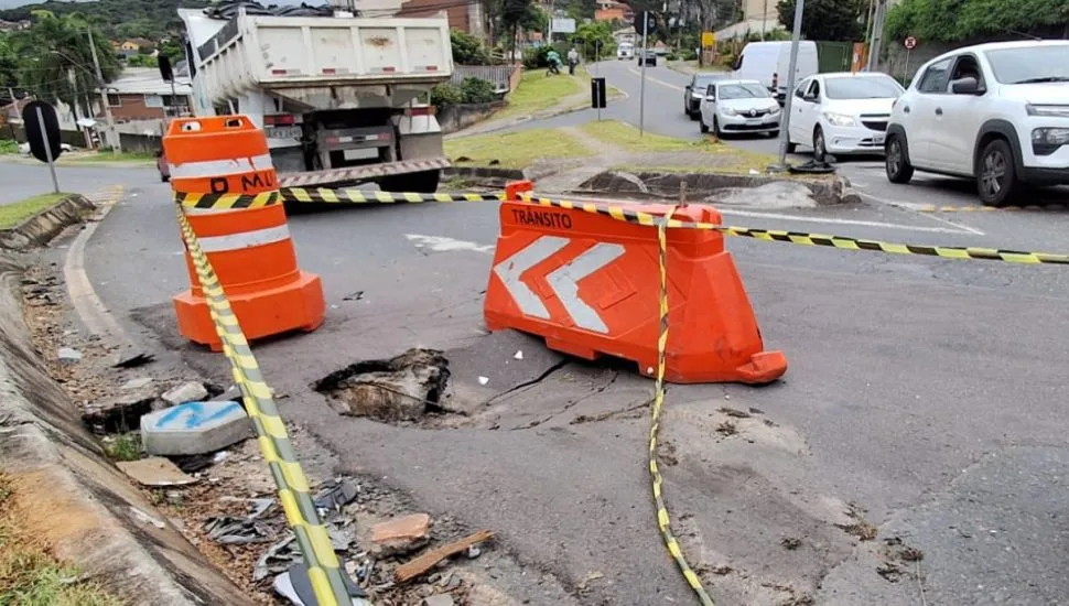 Imagem mostra um buraco na rua, sinalizado com fitas e cavaletes. Ao fundo, veículos no trânsito.