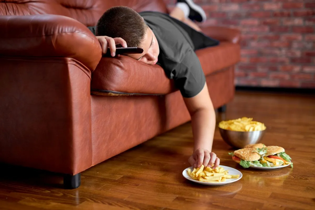 Imagem mostra criança obesa deitada num sofé comendo hamburgueres e batatas fritas que estão em pratos dispostos no chão à sua frente enquanto assiste televisão