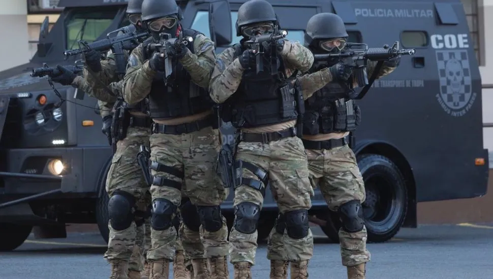 Na imagem, policiais fardados e com armas nas mãos, em frente a um veículo blindado do BOPE.