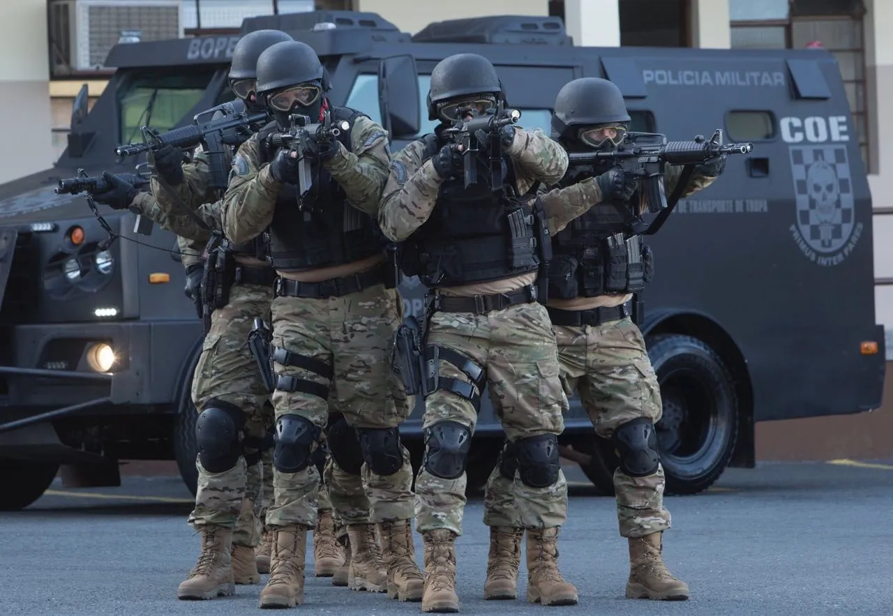 Na imagem, policiais fardados e com armas nas mãos, em frente a um veículo blindado do BOPE.