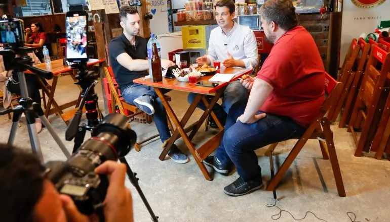 Imagem mostra o candidato Eduardo Pimentel sentado à uma mesa de boteco ao lado dos jornalistas Eduardo Klisiewicz e Leonardo Coleto. O ambiente é um bar cheio de coisas penduradas na perede