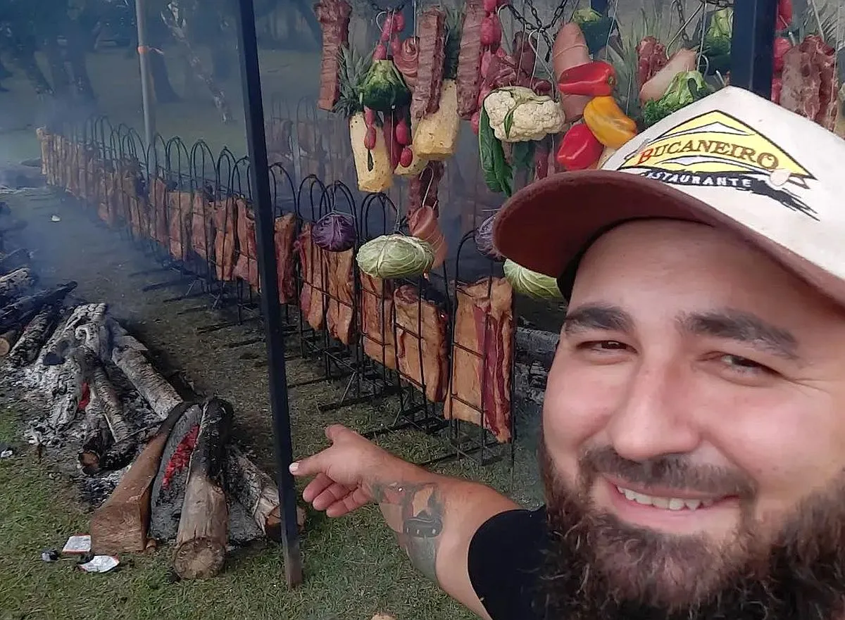 Imagem mostra o chefe marcos oliveira apontando para uma série de costelas sendo assadas em fogo de chão, além de um varal de legumes também sobre as brasas