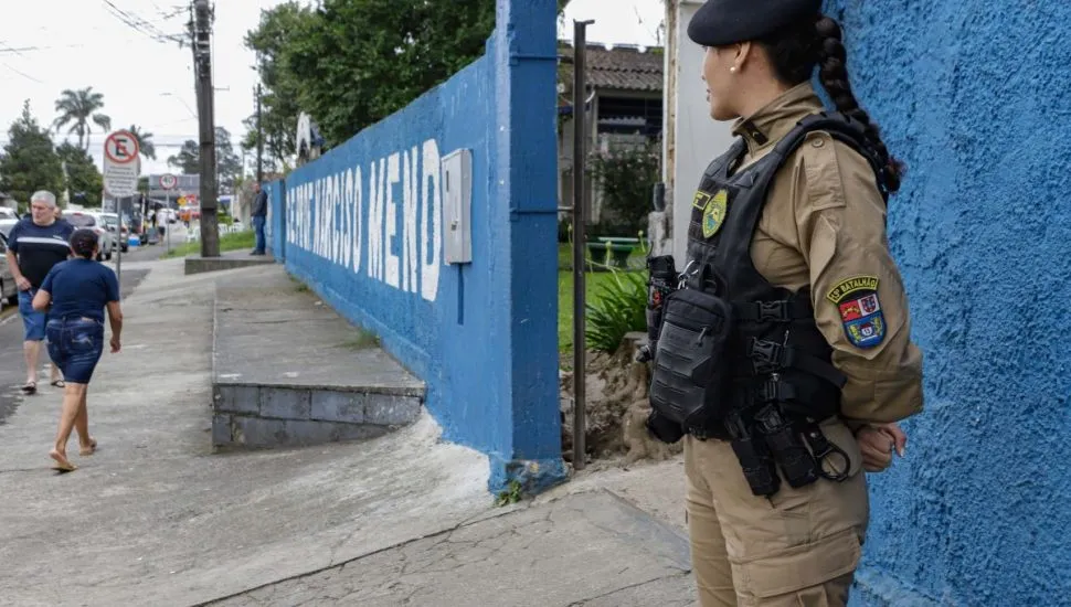 Imagem mostra policial militar na frente de uma escola, enquanto eleitores passam pela calçada