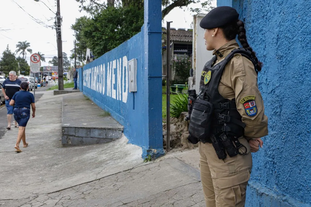 Imagem mostra policial militar na frente de uma escola, enquanto eleitores passam pela calçada