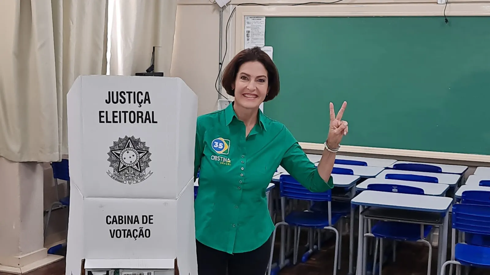 Imagem mostra candidata Cristina Graeml em uma sala de escola, com carteiras e um quadro negro ao fundo. Ela está na frente da Cabina de Votação, fazendo um gesto de V da Vitória após votar