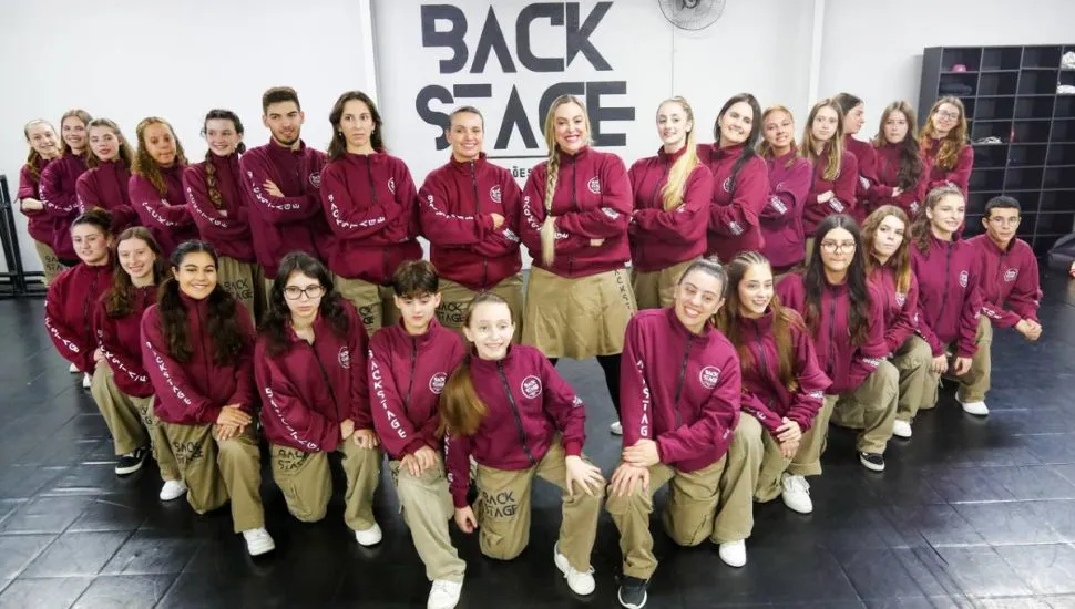 Imagem mostra integrantes de uma escola de dança de Curitiba. Eles estão numa formação em V com blusa bordô e calça bege.