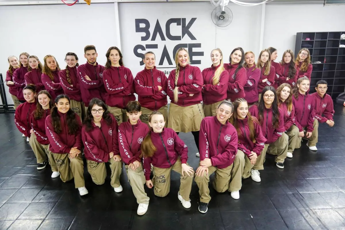 Imagem mostra integrantes de uma escola de dança de Curitiba. Eles estão numa formação em V com blusa bordô e calça bege.