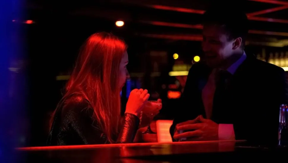 Imagem mostra uma mulher em um bar a noite conversando com um homem de terno.