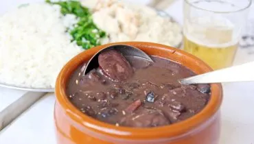 Imagem mostra uma cumbua de feijoada, com um copo de cerveja e um prato de arroz ao lado