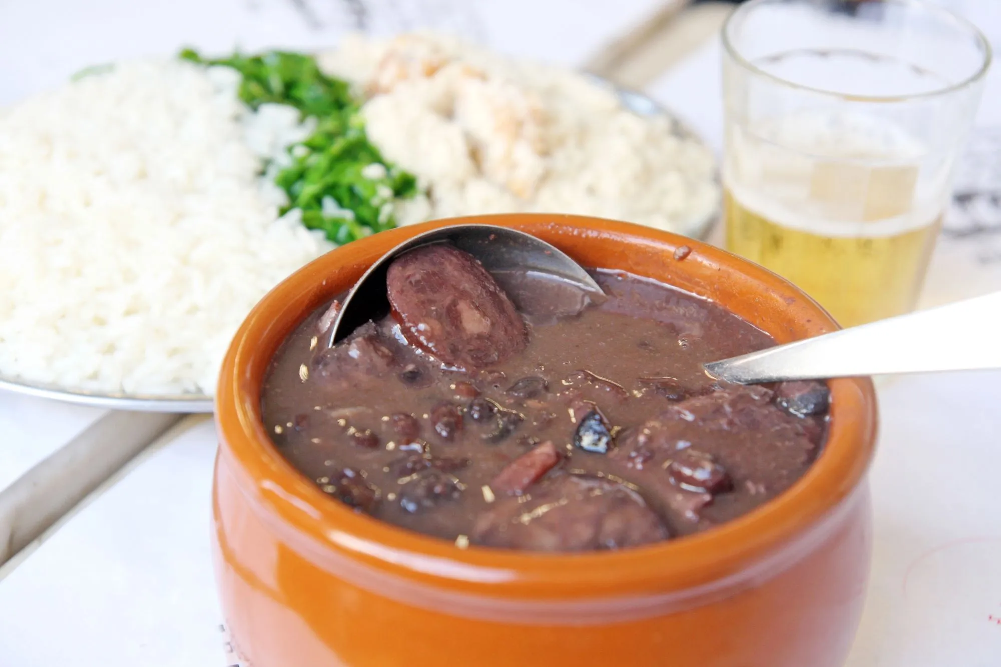 Imagem mostra uma cumbua de feijoada, com um copo de cerveja e um prato de arroz ao lado