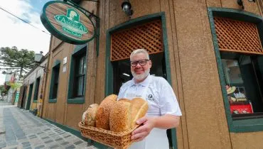 Na imagem, Eduardo Henrique Engelhardt segura cesto com pães na porta da Padaria América, em Curitiba