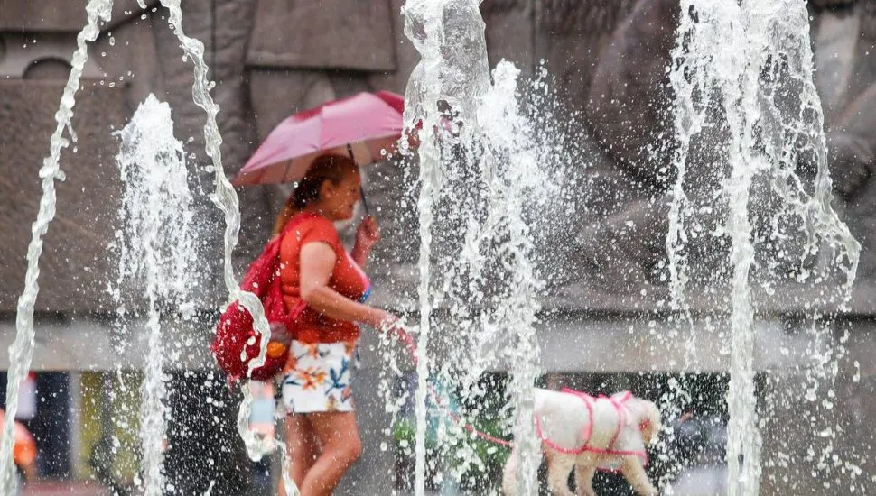 Imagem mostra uma mulher andando com um guarda chuva e um cachorro. Ela está ao fundo e, na frente, jatos de um chafariz.