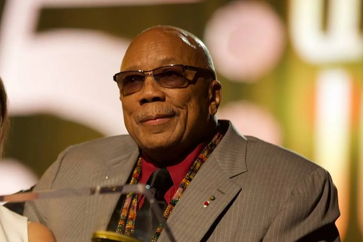 Imagem mostra Quincy Jones, com terno bege, camisa vermelha, óculos escuros e gravata preta.