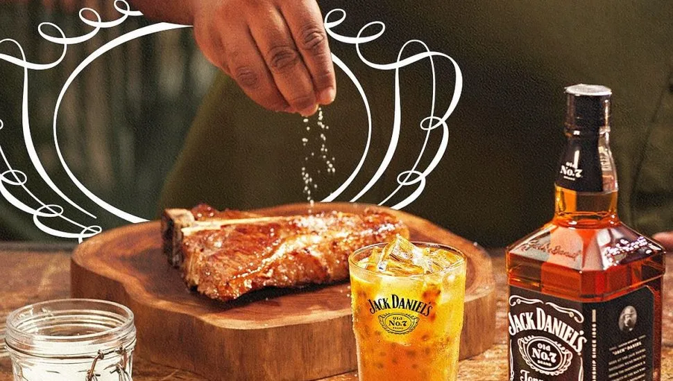 Imagem mostra uma mão salpicando sal granulado em um filé de boi, com um cpo de drink feito com whisky da Jack Daniel's