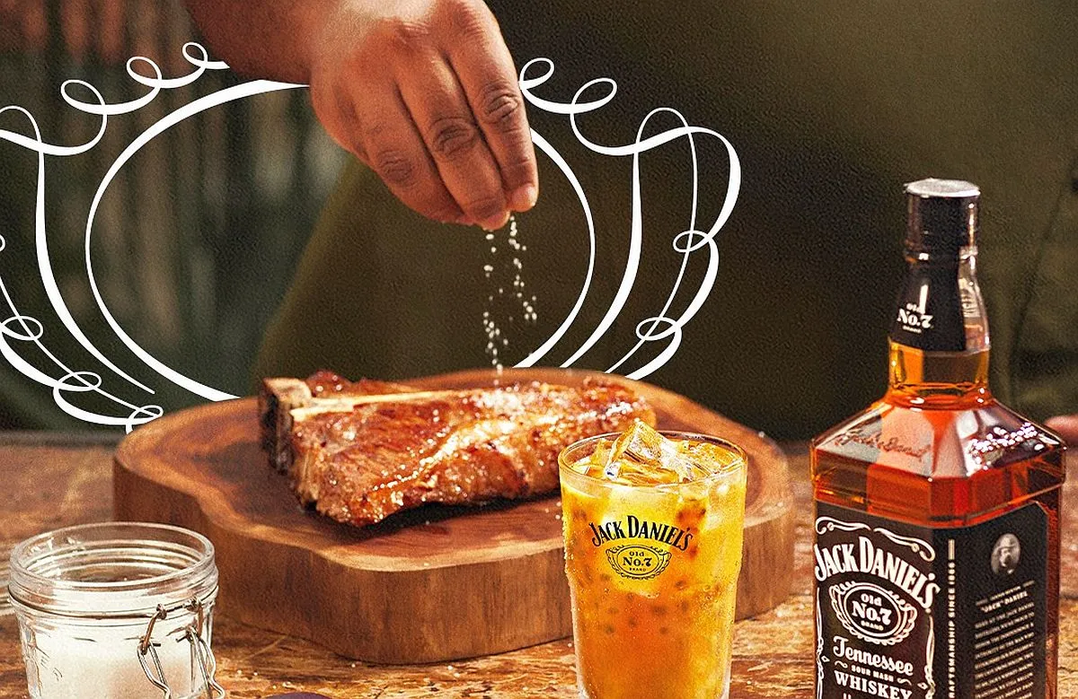 Imagem mostra uma mão salpicando sal granulado em um filé de boi, com um cpo de drink feito com whisky da Jack Daniel's