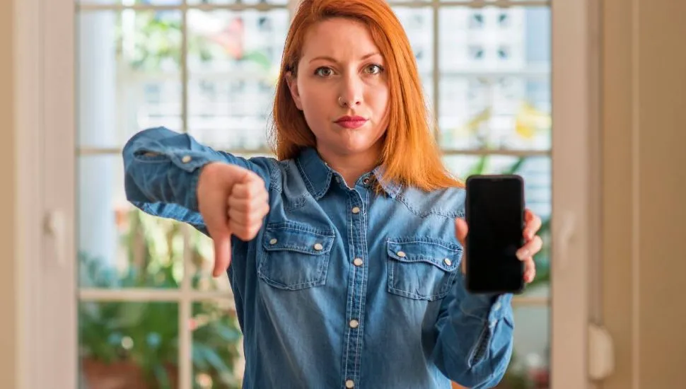 Imagem mostra uma mulher ruiva segurando um celular e fazendo um joinha pra baixo.