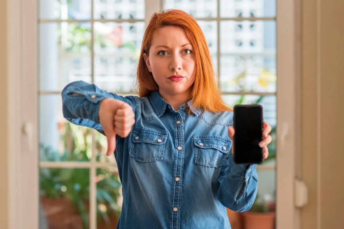 Imagem mostra uma mulher ruiva segurando um celular e fazendo um joinha pra baixo.