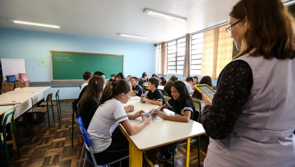 Na imagem, uma professora de costas dá aula em uma sala com alunos sentados, que dividem grandes mesas.